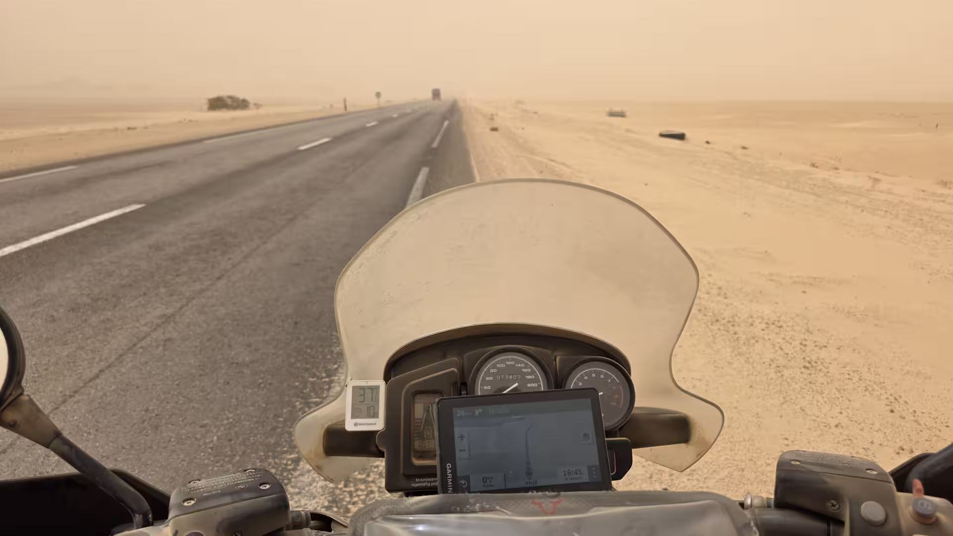 Blick über Lenker und Windschild eines Motorrads auf eine gerade Straße durch eine karge, staubige Landschaft.