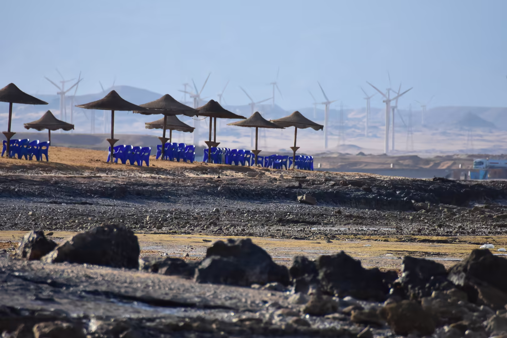 Blaue Stühle unter Strohdächern am Roten Meer, dahinter der Windpark.
