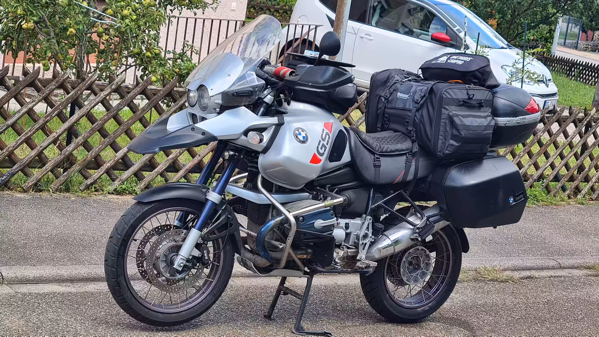 Ein voll beladenes BMW-Reise-Motorrad mit Packtaschen steht am Rand einer Straße vor einem Holzgitterzaun.
