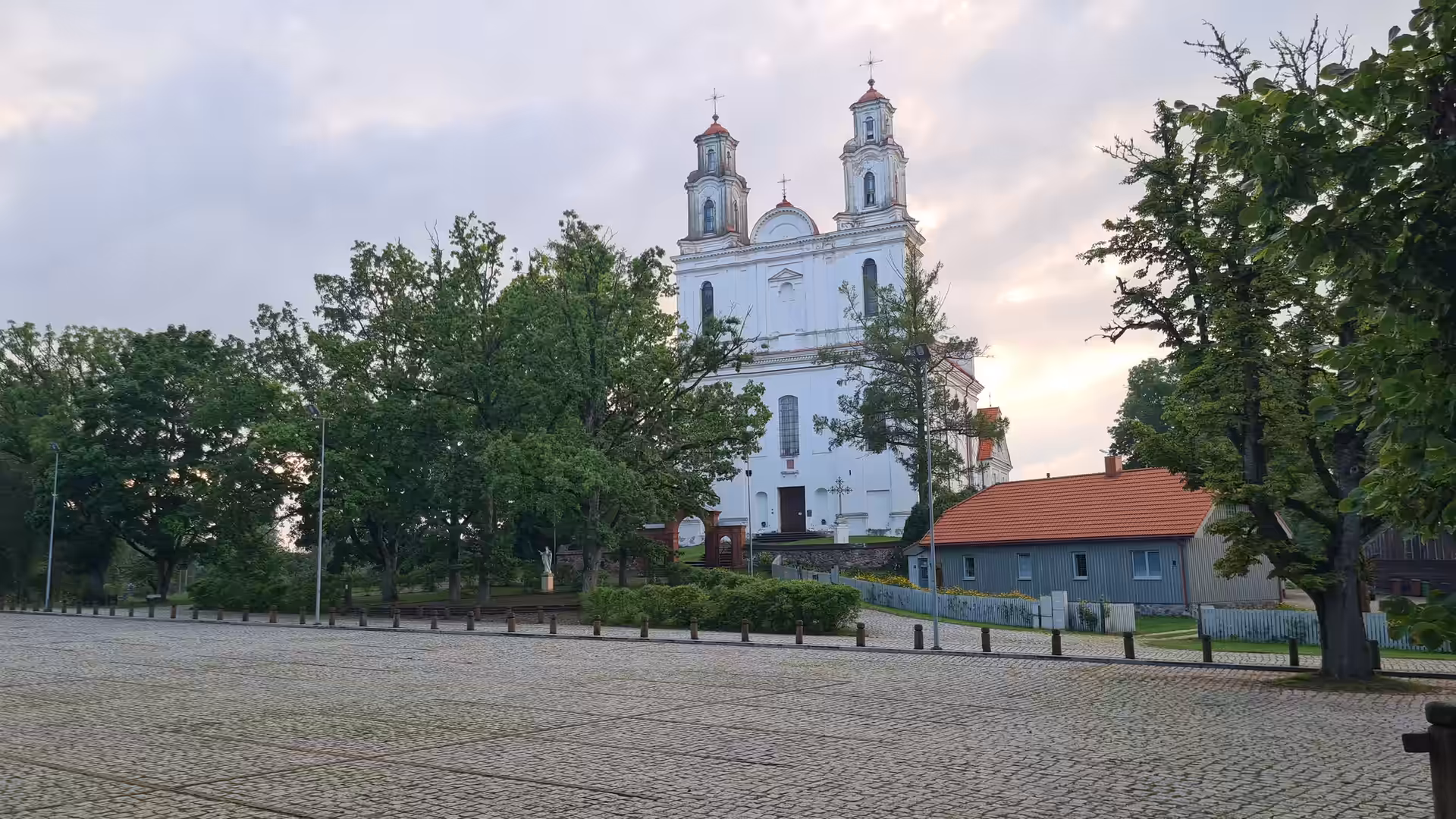 Weiße Kirche mit zwei Türmen und roten Dächern über einem gepflasterten Dorfplatz.
