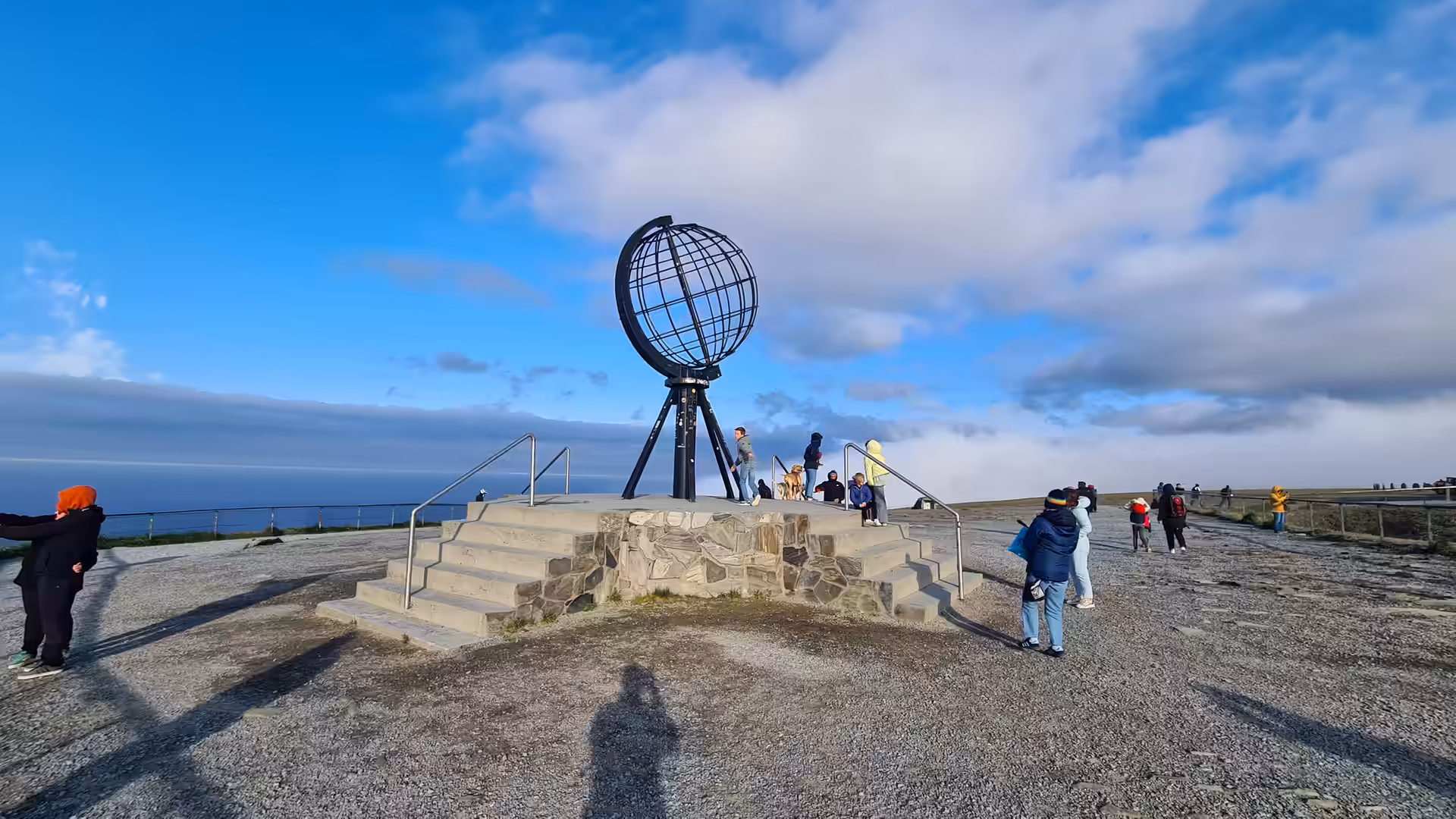 Besucher stehen um eine kugelförmige Metallgitter-Skulptur auf einer kargen Felshochfläche vor dem Meer.